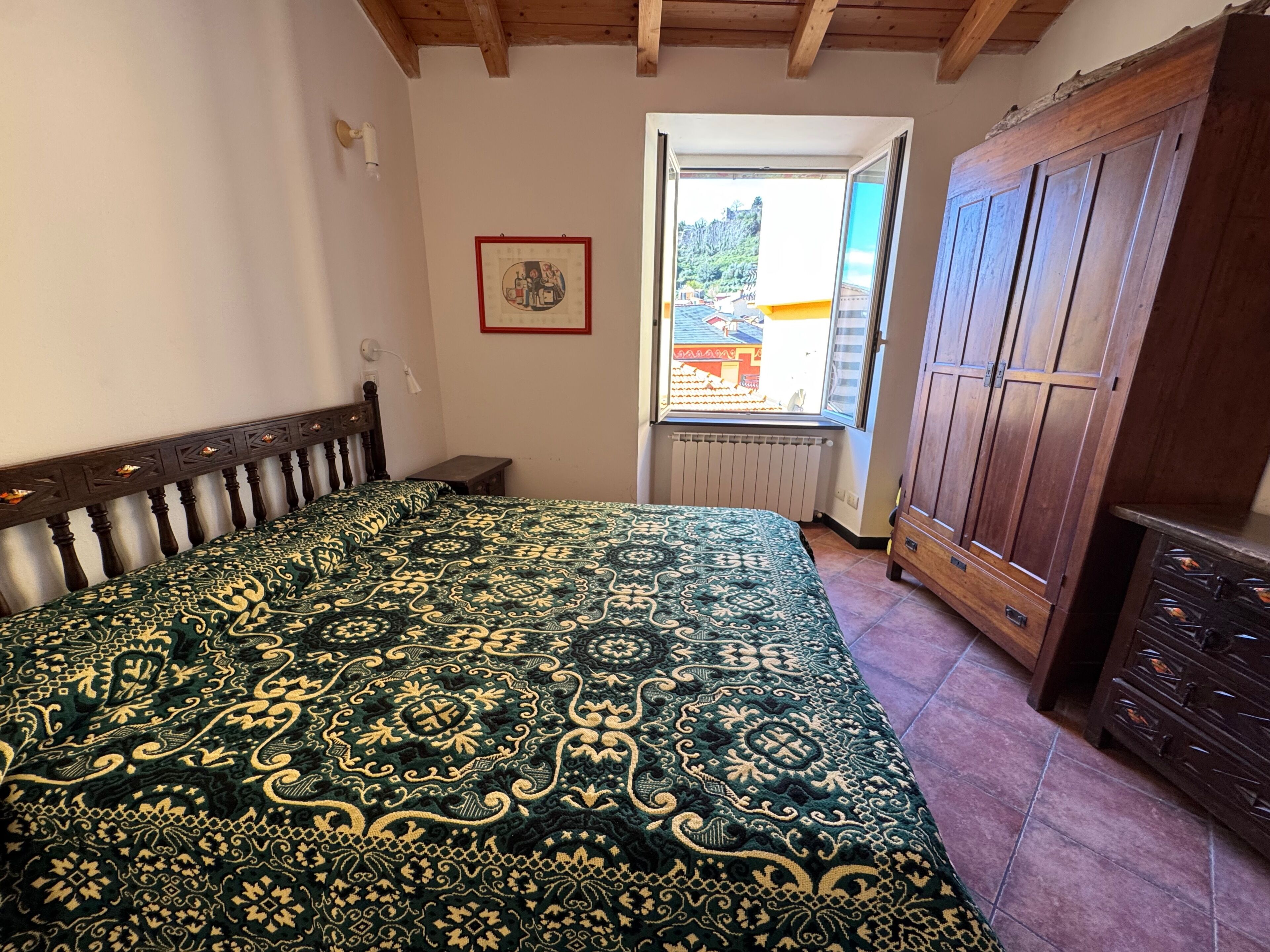 A bedroom with a large bed, a wooden wardrobe, a small bedside table, a radiator, and a window with a view of buildings and trees.
