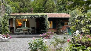 A house with a patio area featuring a table and chairs, surrounded by a garden with various plants and flowers.