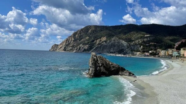 A coastal beach with clear turquoise waters, a rocky outcrop, and a mountain in the background.