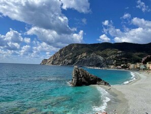 A coastal beach with clear turquoise waters, a rocky outcrop, and a mountain in the background.