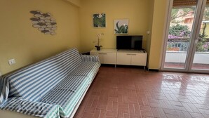 Ein Zimmer mit einer gestreiften Couch, einem Fernseher auf einem Schrank, einem Deckenventilator und einer Glastür zum Außenbereich.