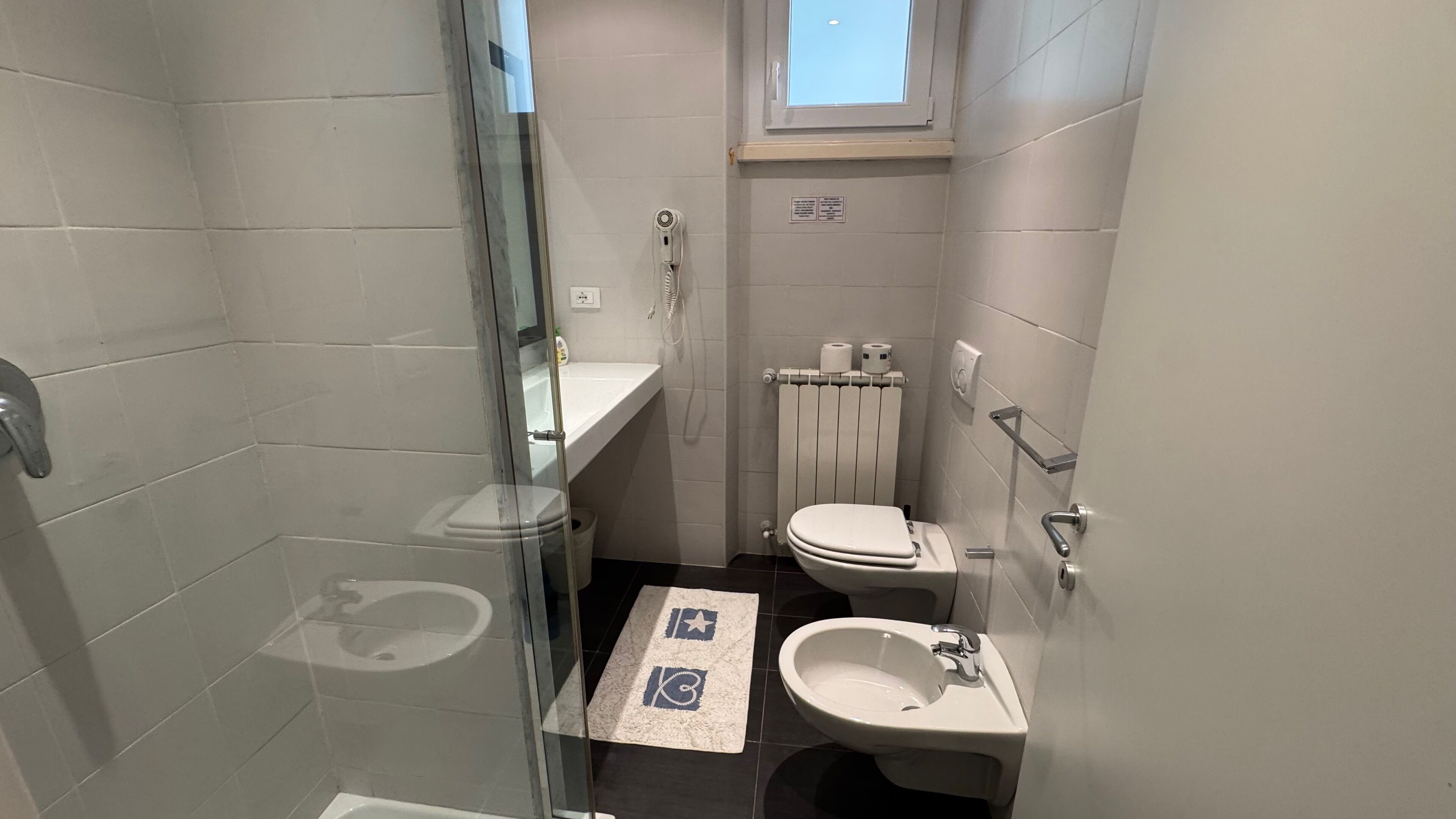 A small, modern bathroom with a glass shower enclosure, a toilet, a sink, and a window above the toilet.