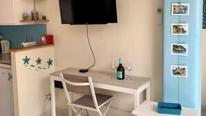 Une pièce de taille modeste, dotée d’un téléviseur à écran plat fixé au mur, d’un petit bureau avec une chaise et d’une étagère ornée d’objets décoratifs.