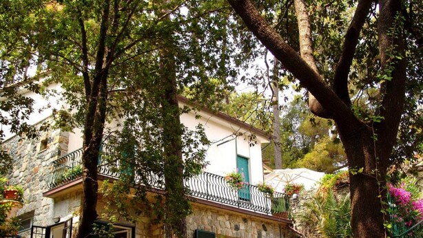 Edificio de piedra con balcón, rodeado de árboles y plantas.