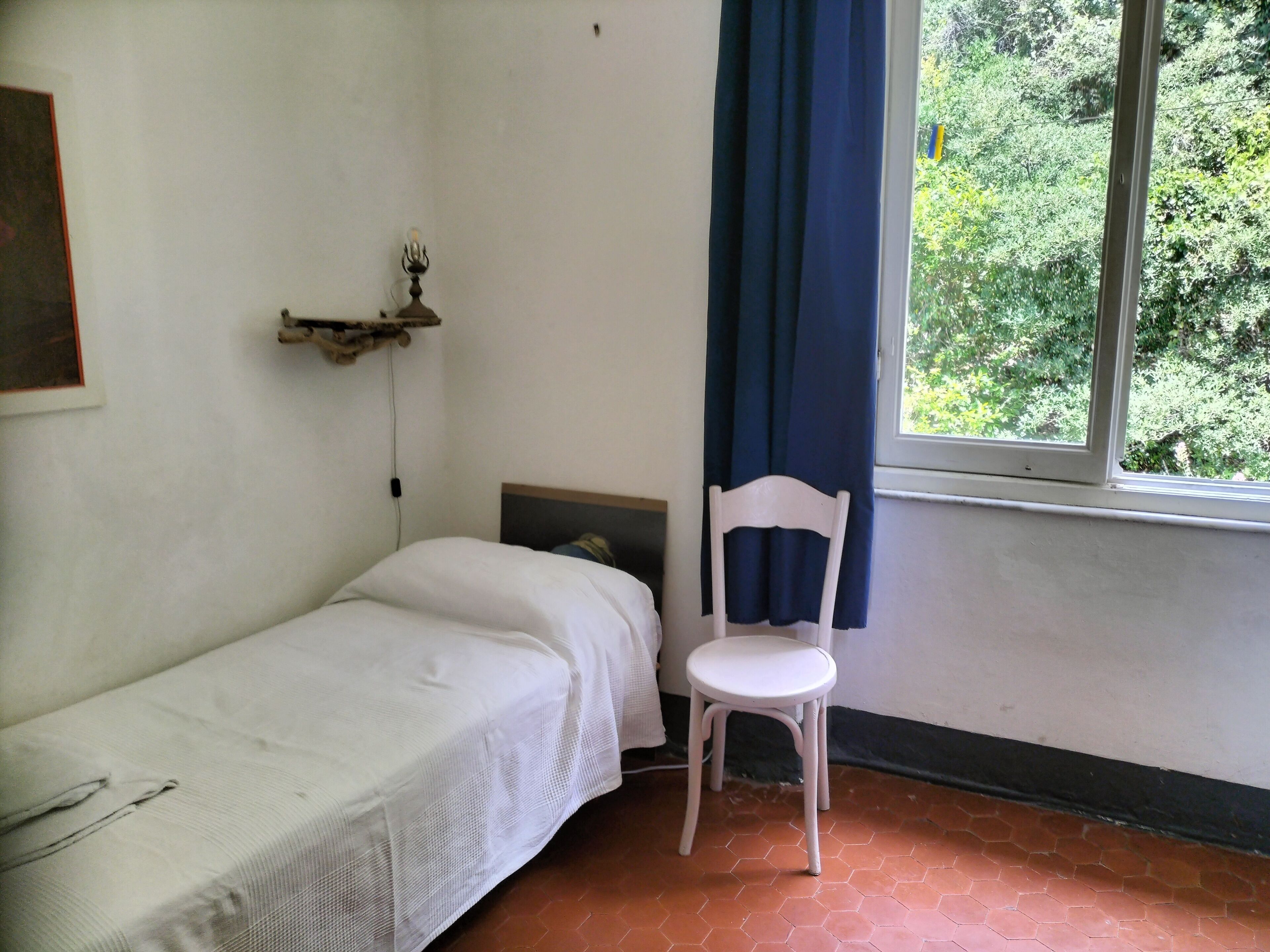 A small, simple room with a single bed, a chair, a window with a view of greenery, and a wall-mounted shelf.
