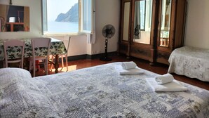 Ein Schlafzimmer mit großem Fenster, einem Holzschrank, zwei Betten, einem Tisch mit Stühlen und einem Ventilator.
