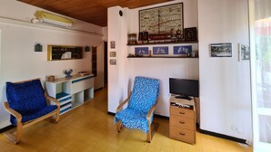Ein Zimmer mit Holzboden, einem blau gemusterten Sessel, einem Fernseher und unterschiedlichen Wanddekorationen.