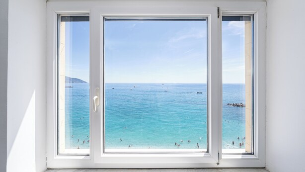 Ein Zimmer mit großem Fenster mit Blick auf ein klares, blaues Meer, Schiffe und Menschen.
