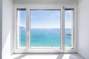 Quarto com uma grande janela dando para um mar azul e límpido, com barcos e pessoas.