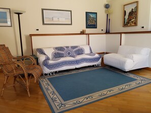 Ein Wohnzimmer mit einem gemusterten Sofa, einem weißen Sessel, einem Korbstuhl und einem blauen Teppich.
