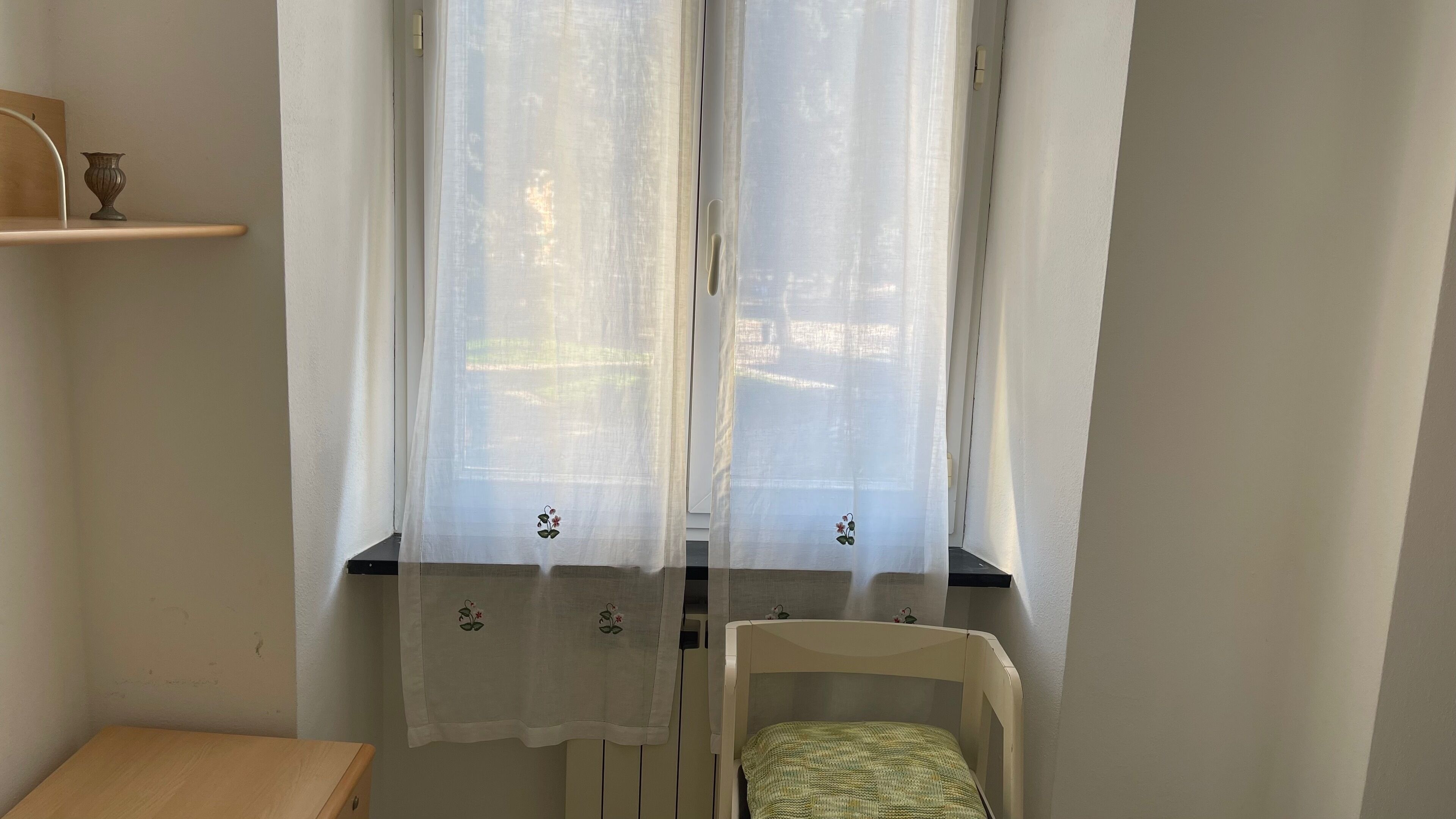 A room with a window covered by sheer curtains, a small table with a vase, and a chair with a green cushion.