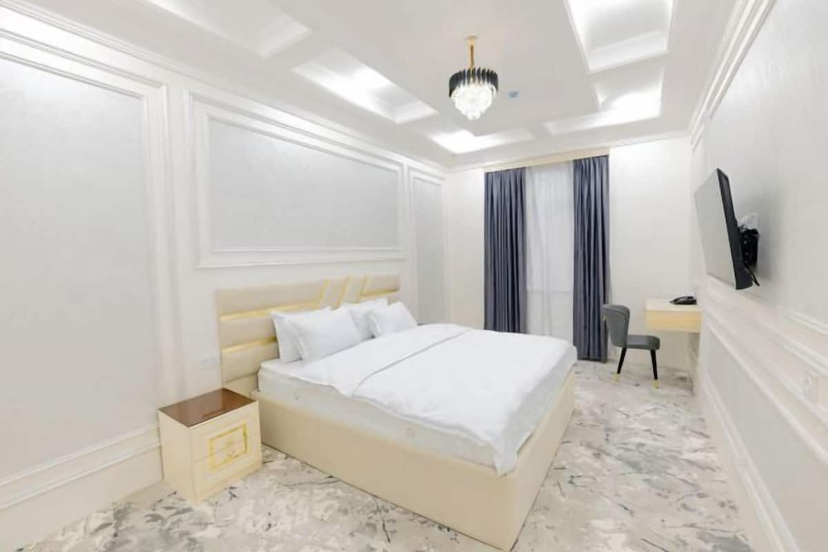 A bedroom with a bed, bedside table, chair, desk, and a TV on the wall.