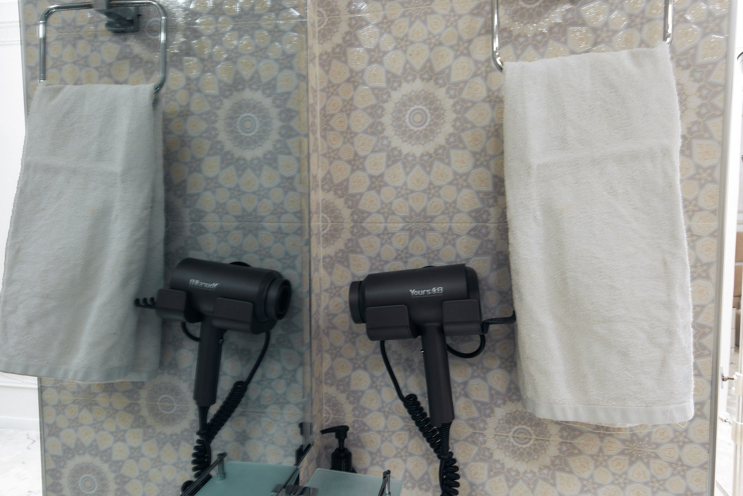 A bathroom with two hair dryers mounted on the wall, a towel rack with two white towels, and a patterned tile backsplash.