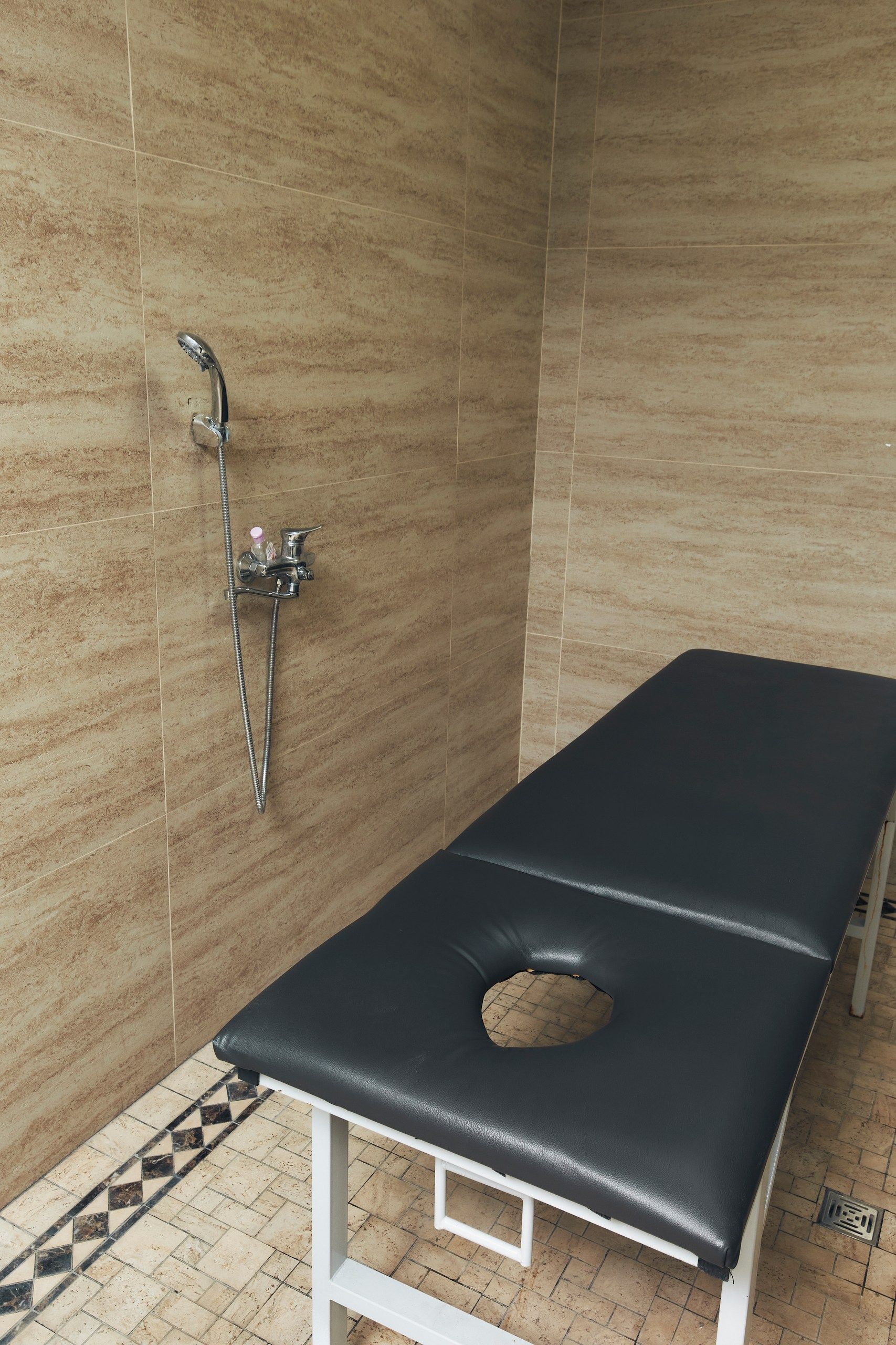 Quarto com mesa de massagem preta, área de chuveiro com chuveiro manual e paredes revestidas com azulejos.