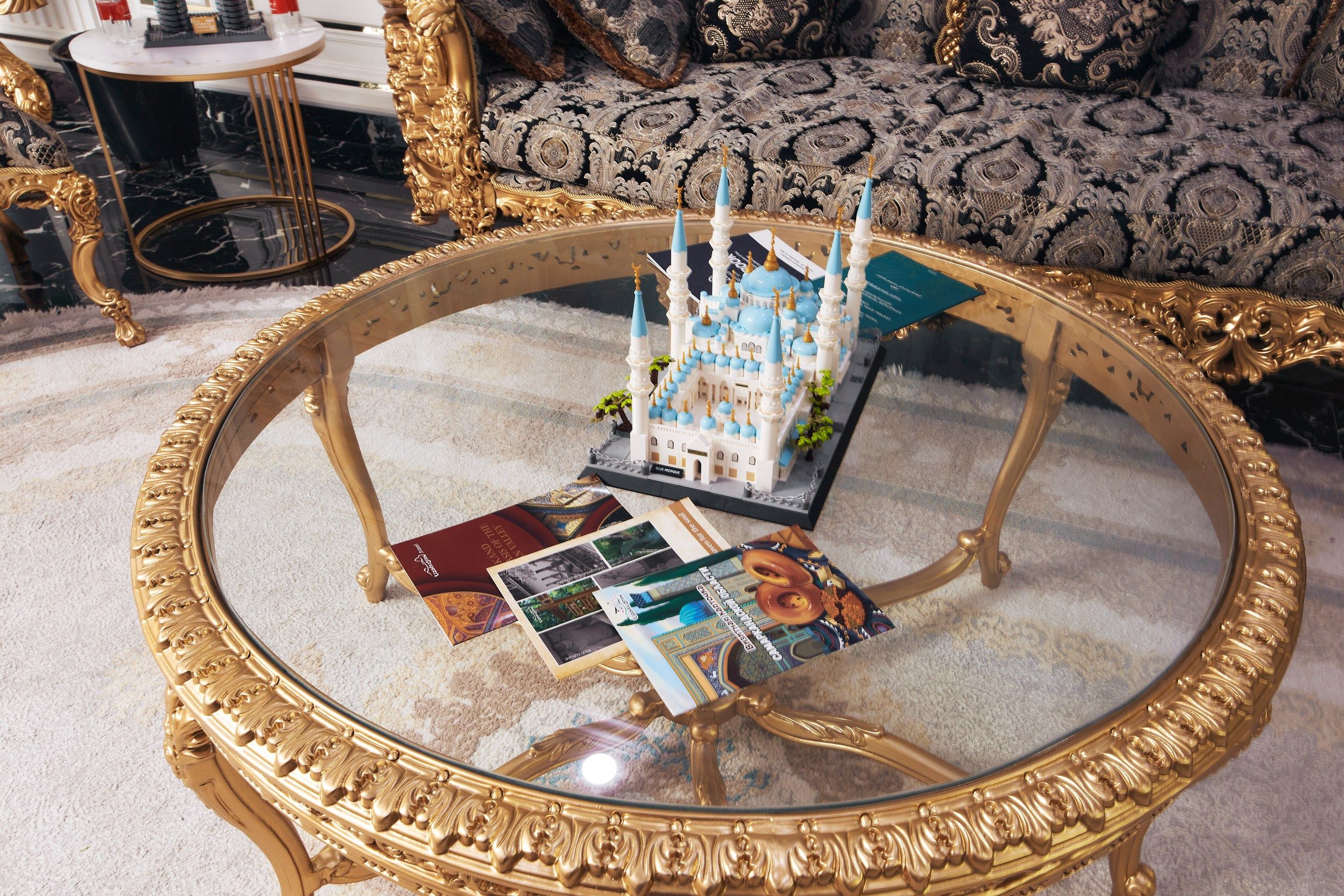 Une table basse ronde en verre avec un cadre doré, une maquette miniature d’une mosquée et plusieurs brochures.