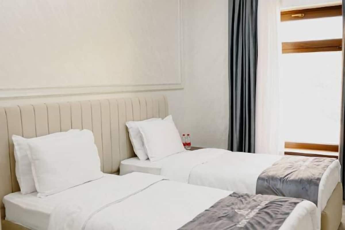 A hotel room with two beds, a nightstand, a window with curtains, and a radiator.