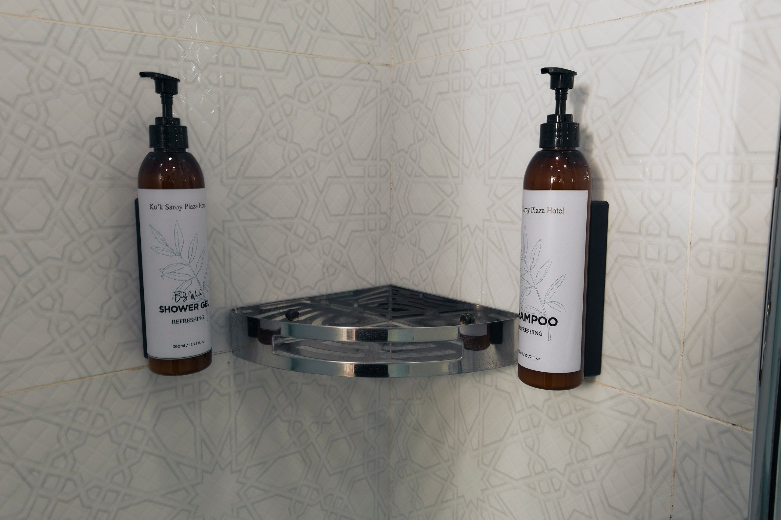 Deux bouteilles de gel douche et de shampoing sont posées sur un plateau dans la douche, dans une salle de bain dont le carrelage est orné de motifs.
