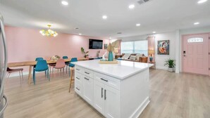Une cuisine moderne avec un îlot central, un coin repas doté de chaises colorées et un mur d’accent rose.