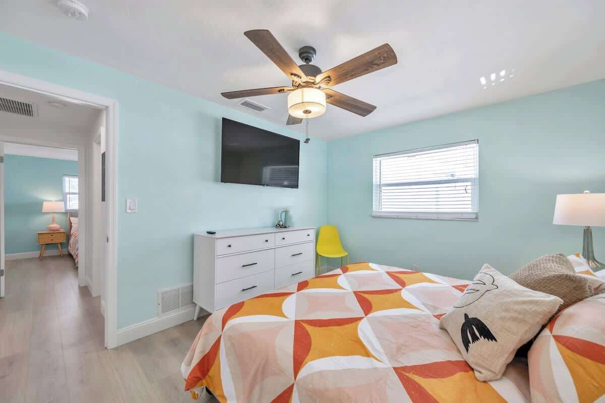 Un dormitorio con cama, ventilador de techo, televisor montado en la pared, ventana con persianas y puerta que da a otra habitación.