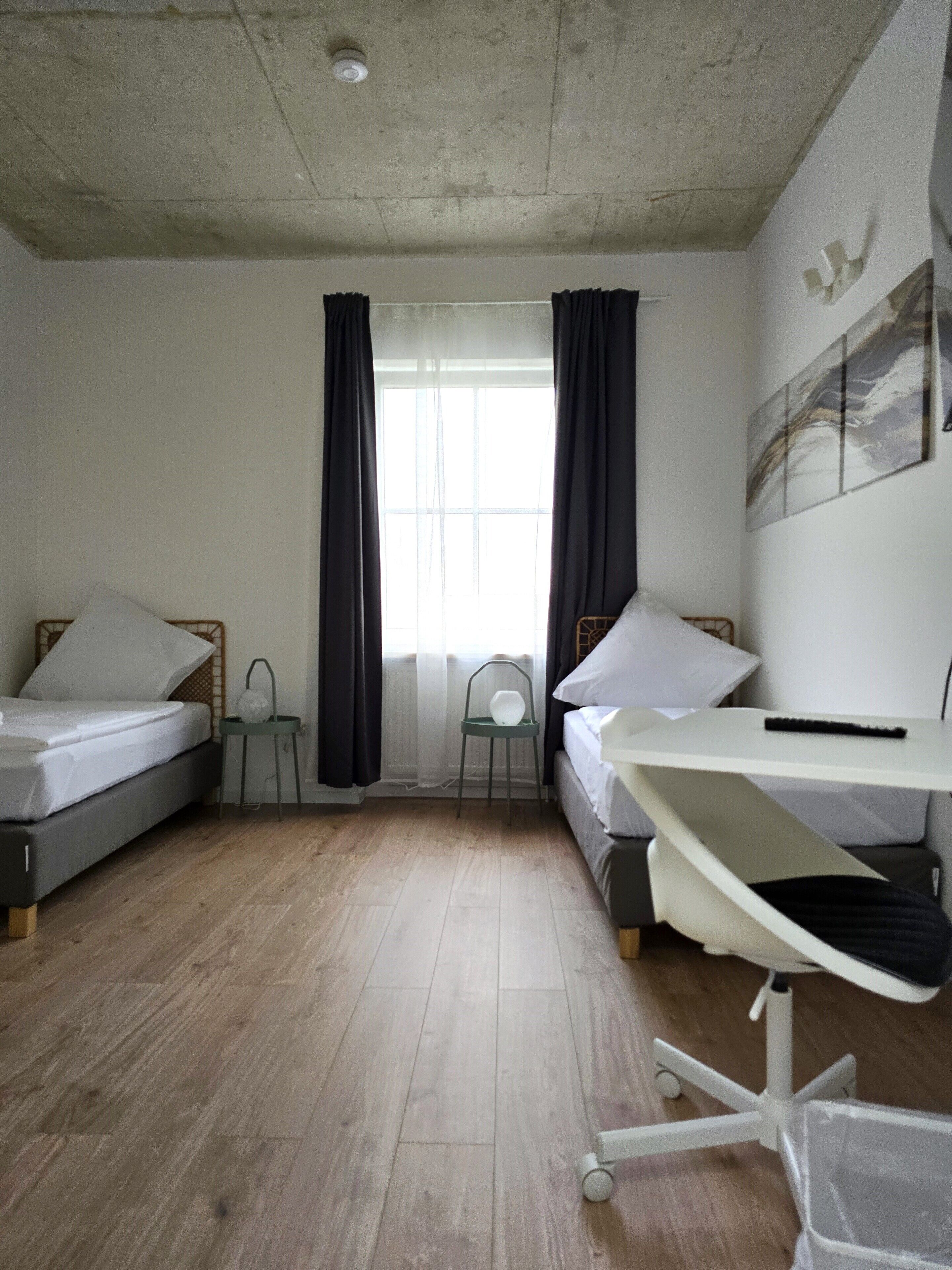 A compact room with two single beds, a desk with a chair, a radiator, and a window with curtains.