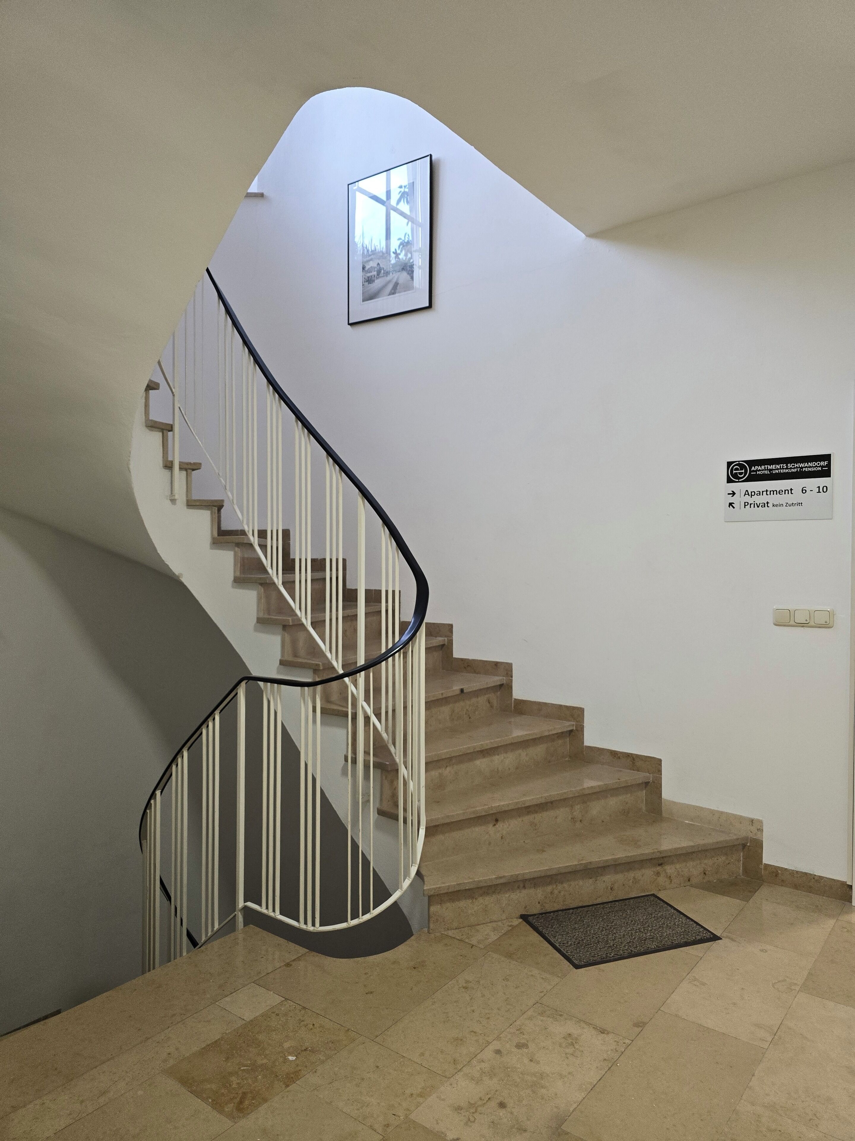 A spiral staircase with white railings and beige steps, leading to a landing with a framed picture and a sign indicating private apartments 6-10.