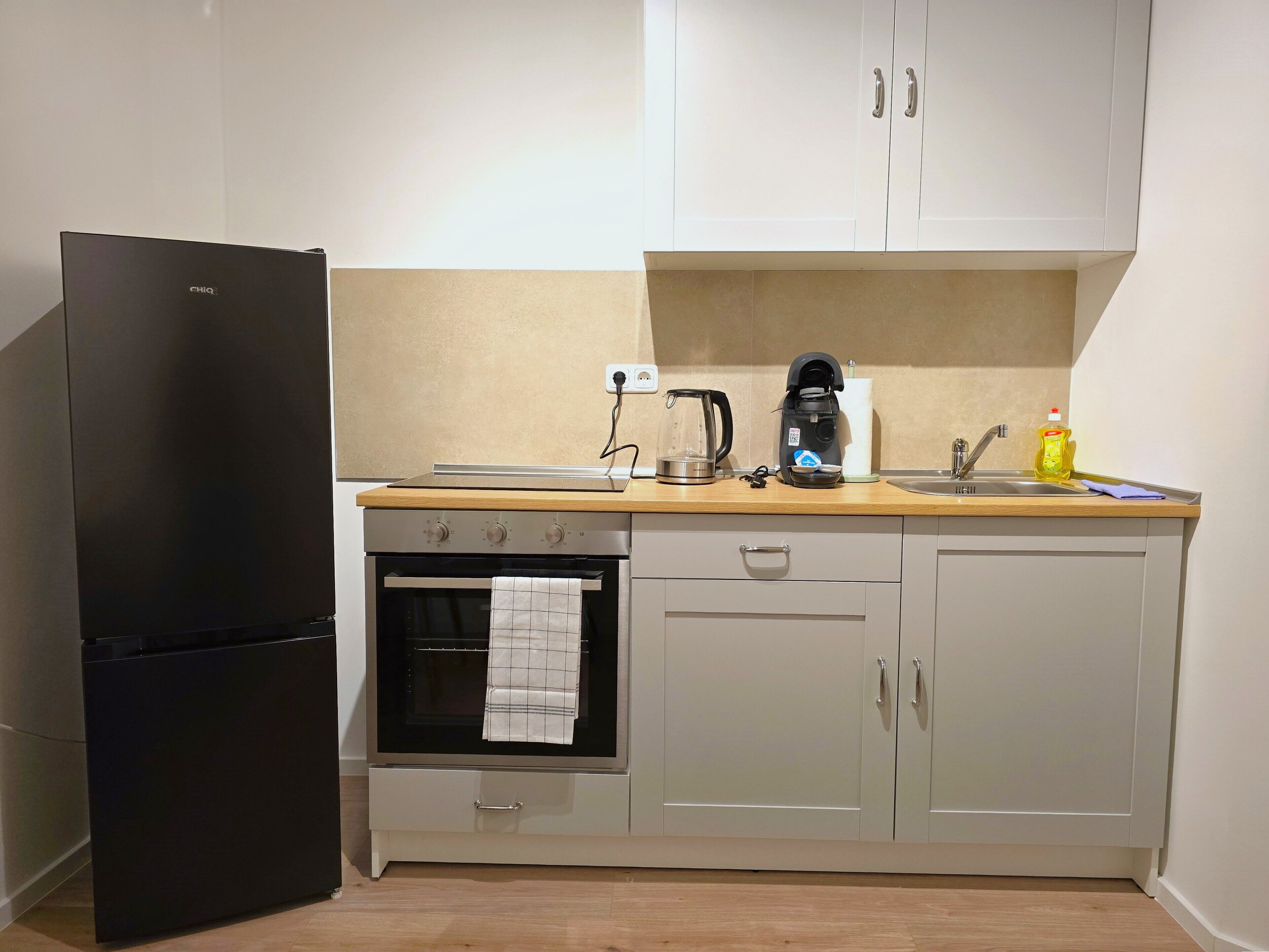 Una cocina compacta con un refrigerador negro, un horno empotrado, una cafetera y un lavamanos.