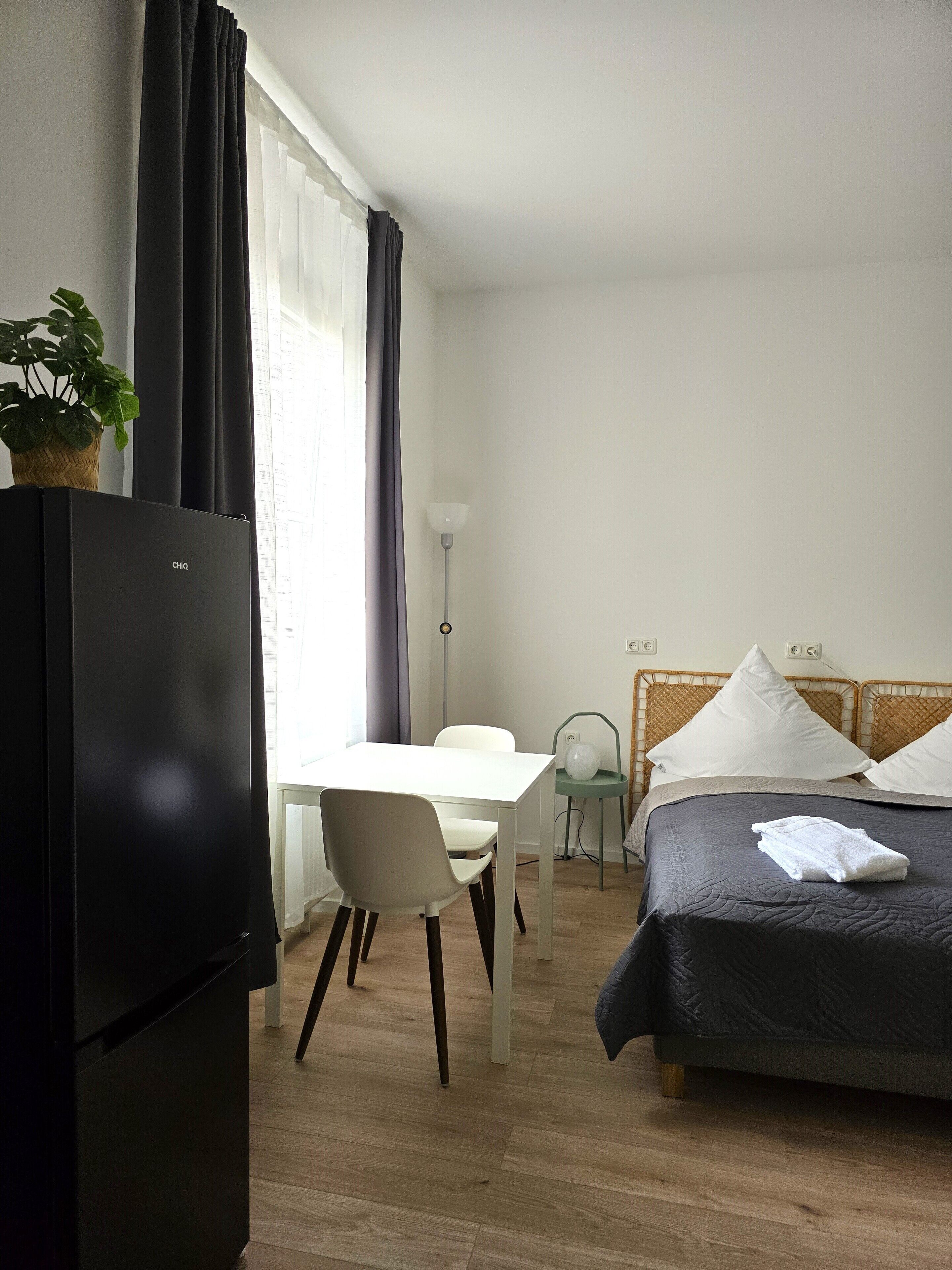 A hotel room with a bed, a desk, a chair, a small table, a lamp, and a refrigerator.