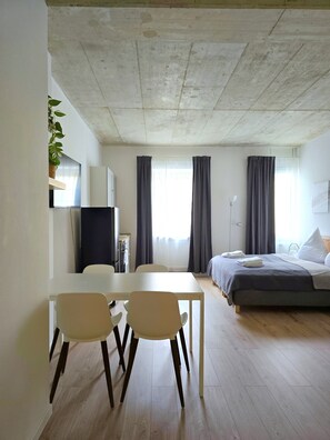 Ein moderner, gut beleuchteter Raum mit einem Bett, einem Esstisch mit Stühlen, einem Fernseher und einer Pflanze.