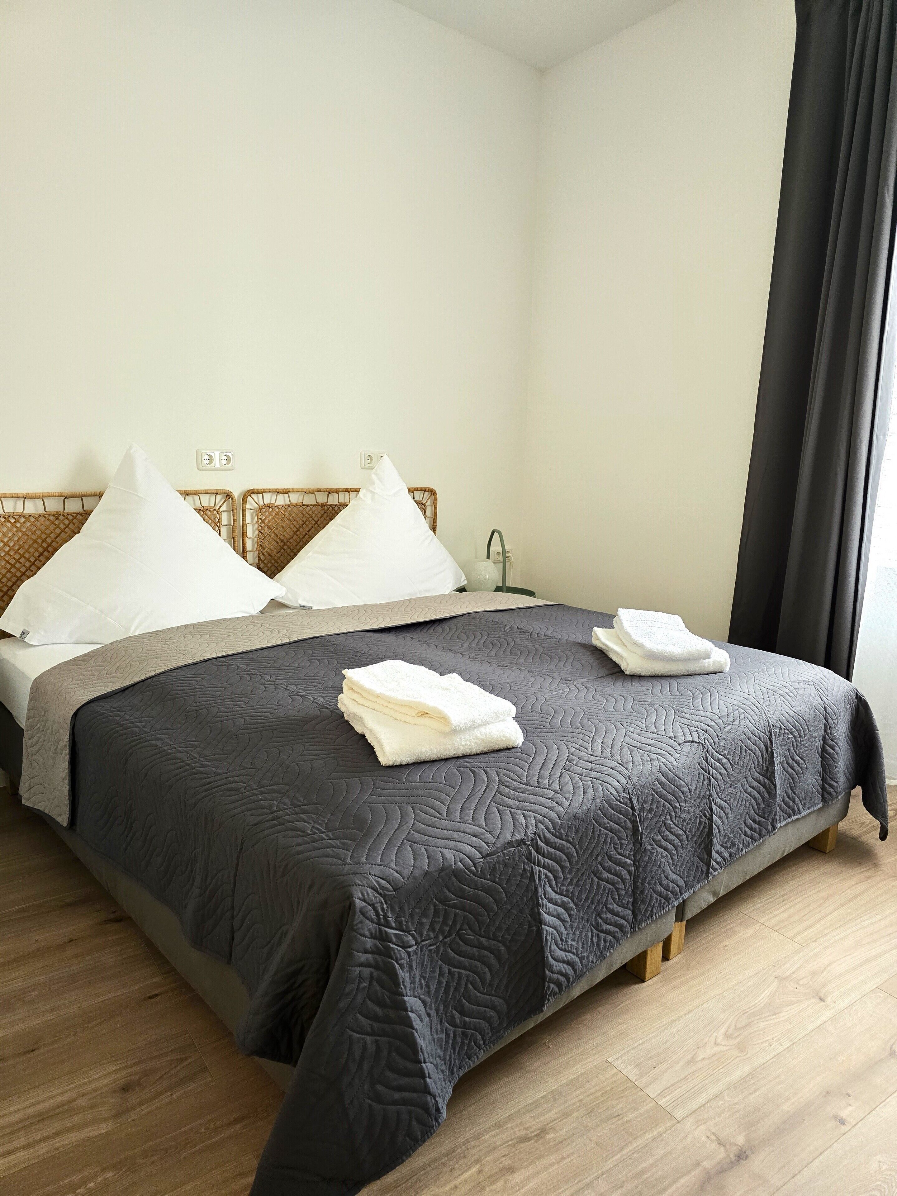A neatly made bed with a dark quilt, white pillows, and folded towels on a wooden floor.