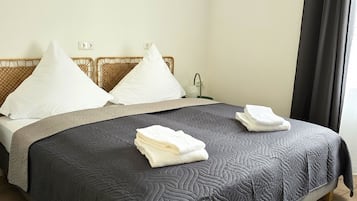 A neatly made bed with a dark quilt, white pillows, and folded towels on a wooden floor.