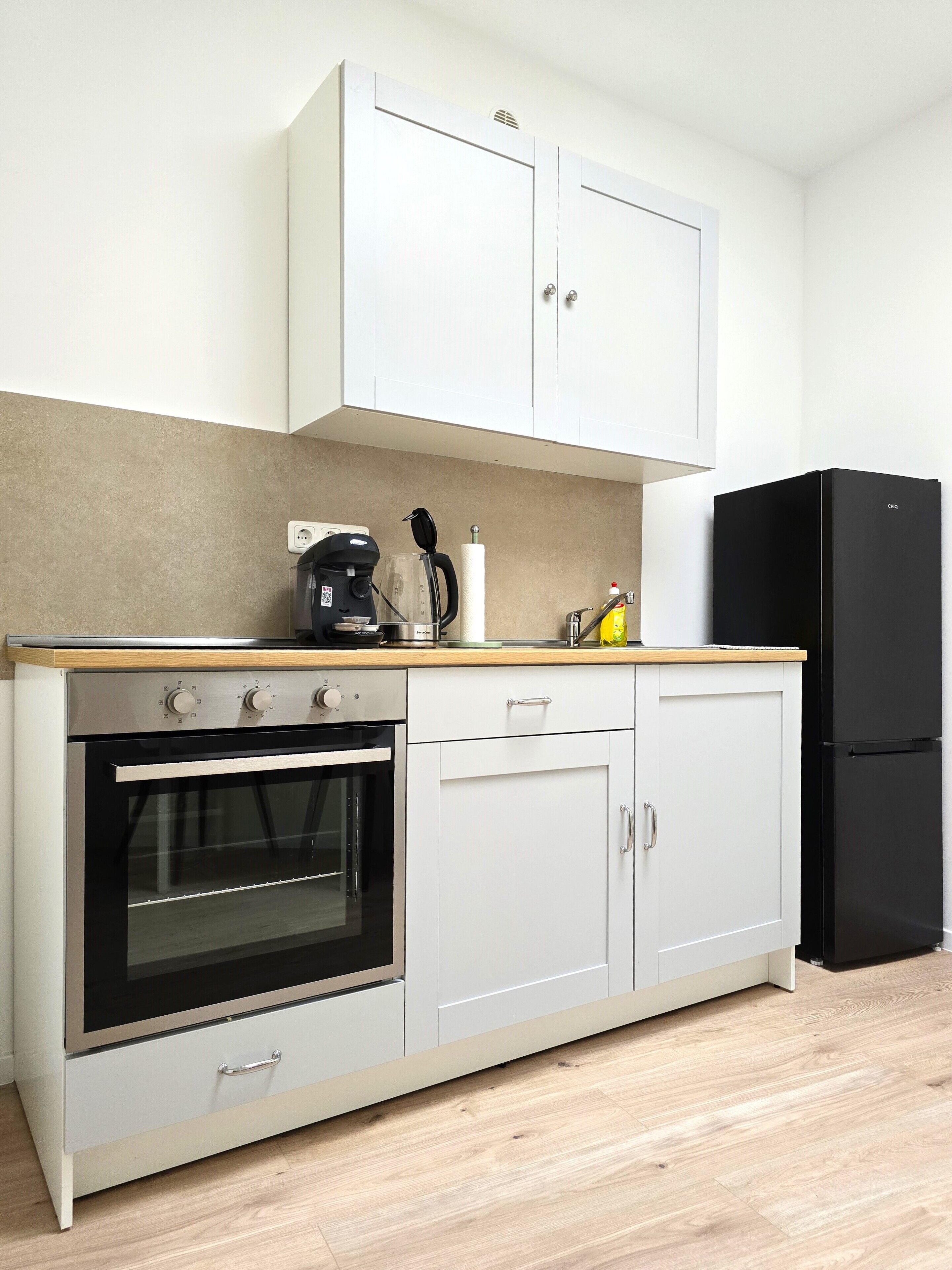 Eine kompakte Küche mit Backofen, Kaffeemaschine und einem schwarzen Kühlschrank.