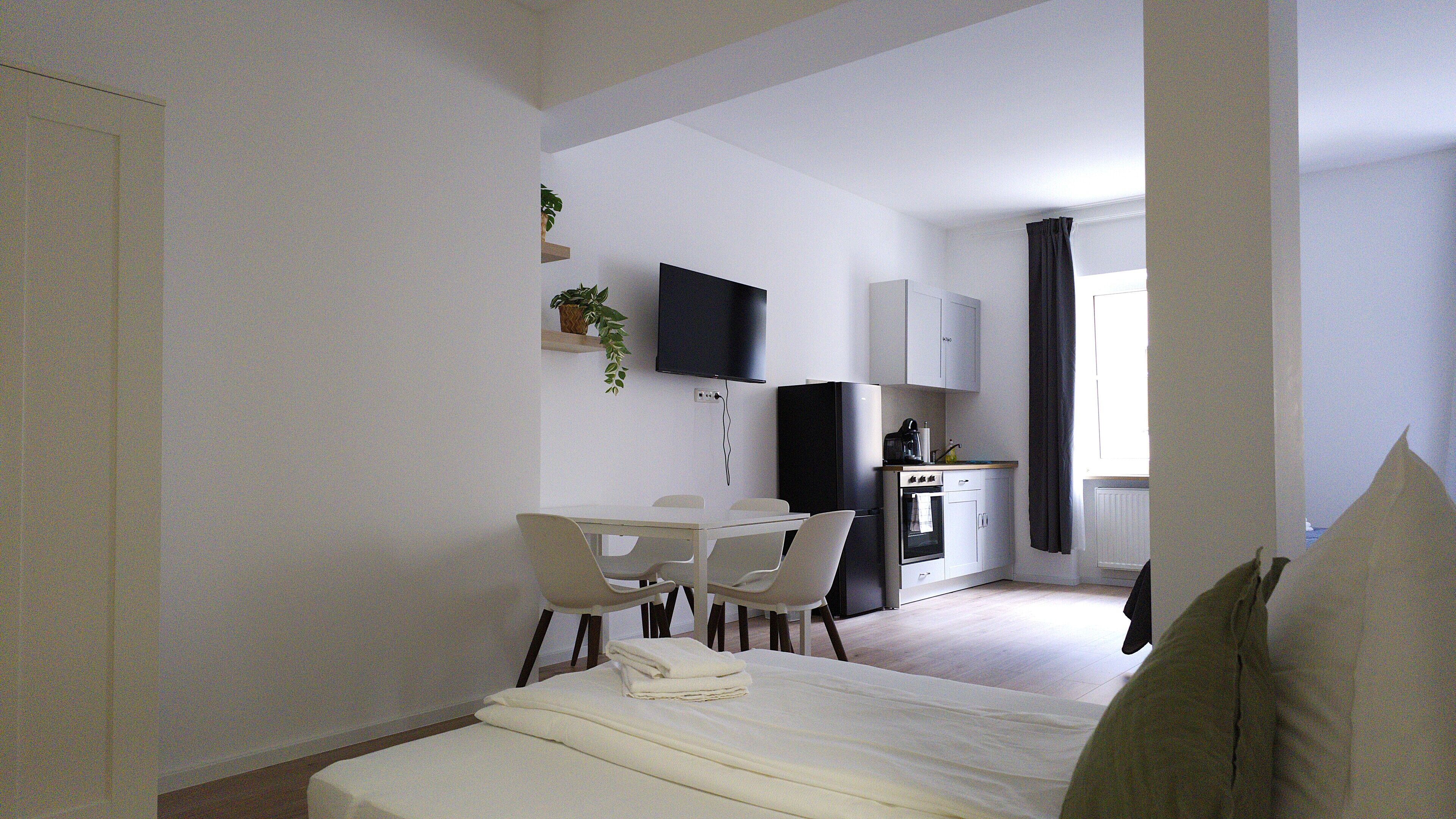 A compact hotel room with a bed, a dining table with chairs, a kitchenette, a TV, and a window with curtains.