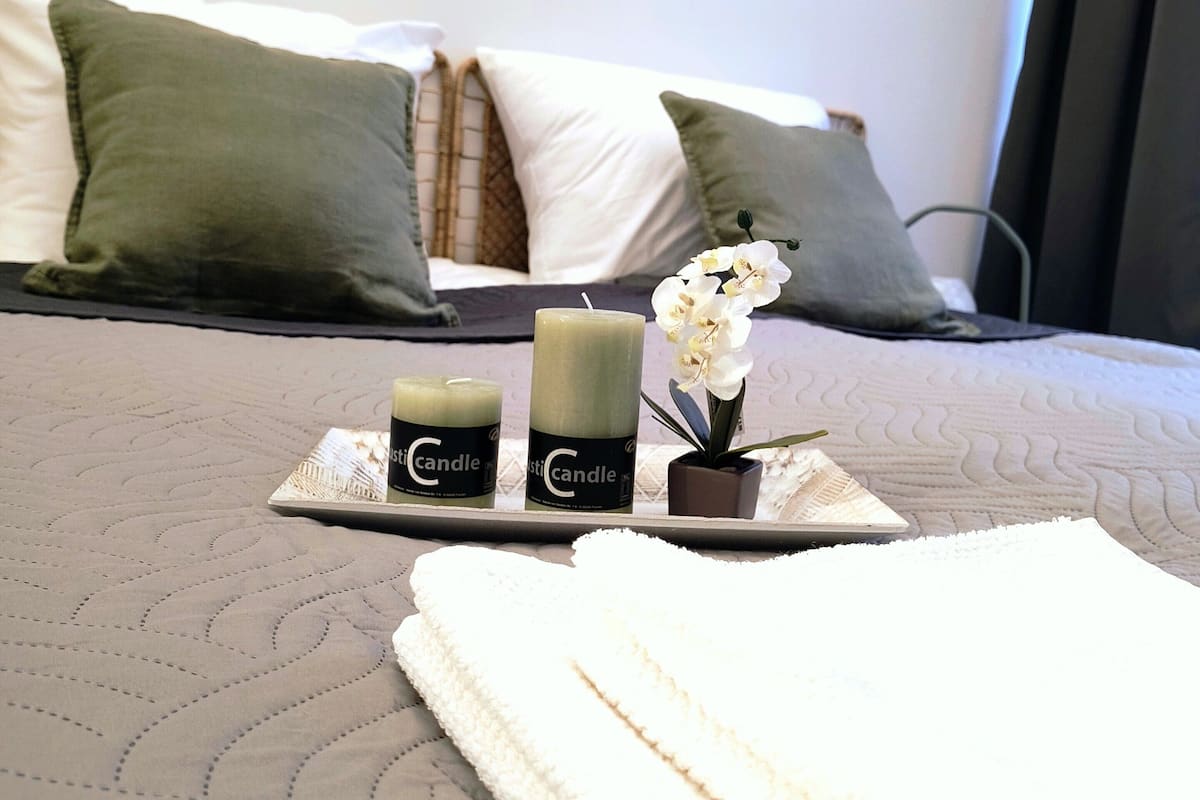 Un lit bien fait, recouvert d’une couette blanche, avec deux bougies, un plateau sur lequel est posée une autre bougie, et une petite orchidée.