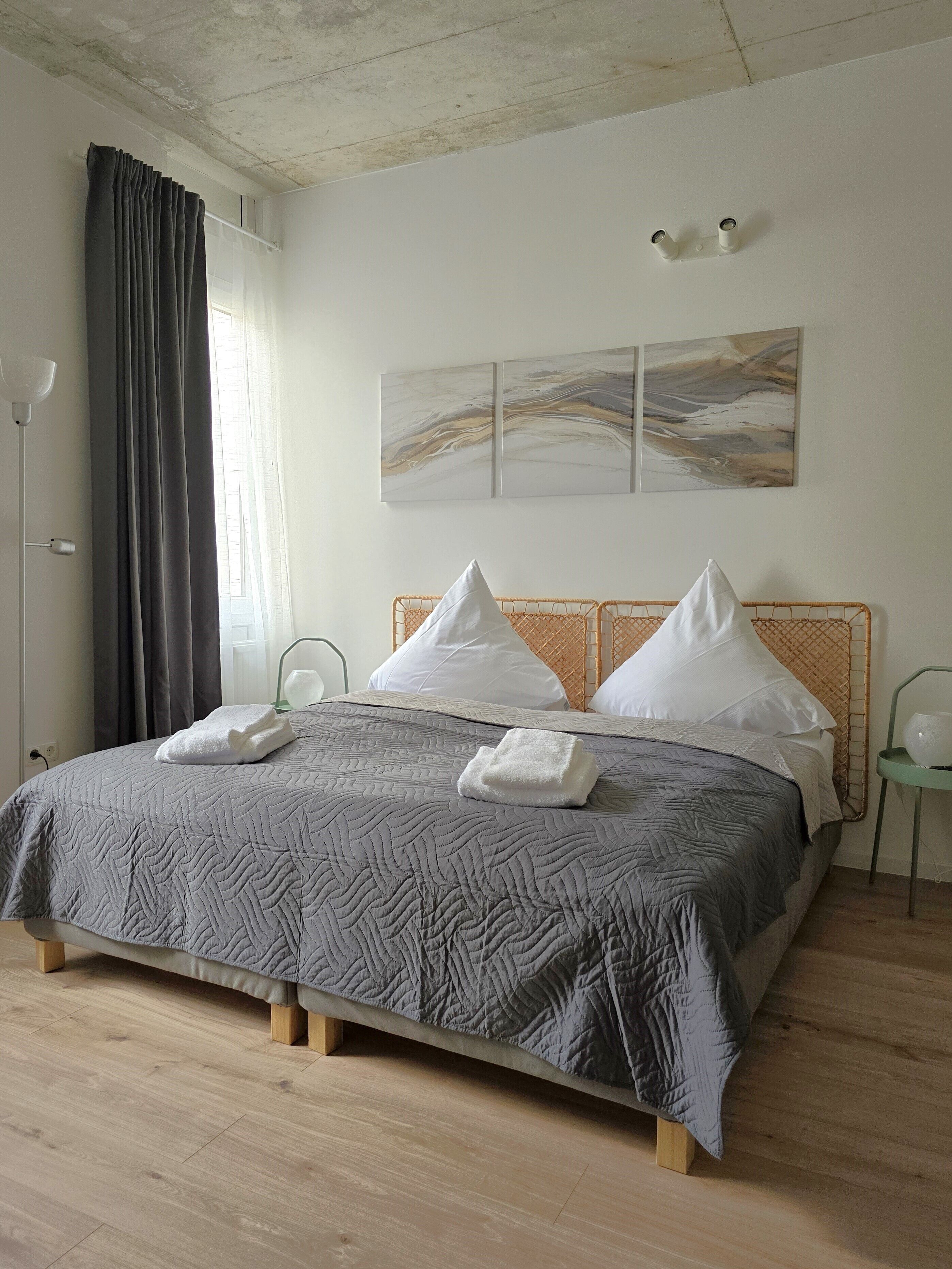 Una cama bien hecha con colcha gris, dos almohadas blancas y dos toallas dobladas. Se aprecia un cabecero de madera, una lámpara de pie y una lámpara de pared.
