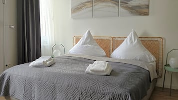 Un lit bien fait, avec une couverture grise, deux oreillers blancs et deux serviettes pliées. On y trouve une tête de lit en bois, une lampe sur pied et un luminaire fixé au mur.
