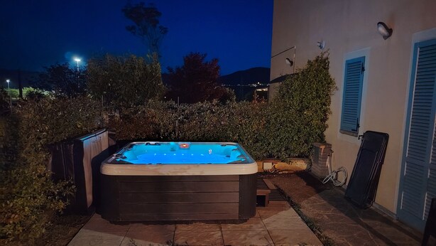 A hot tub in a backyard with a stone patio, surrounded by greenery and a house with blue shutters. - Nociarelle Cottage (Nociarelle-Scarpiello)