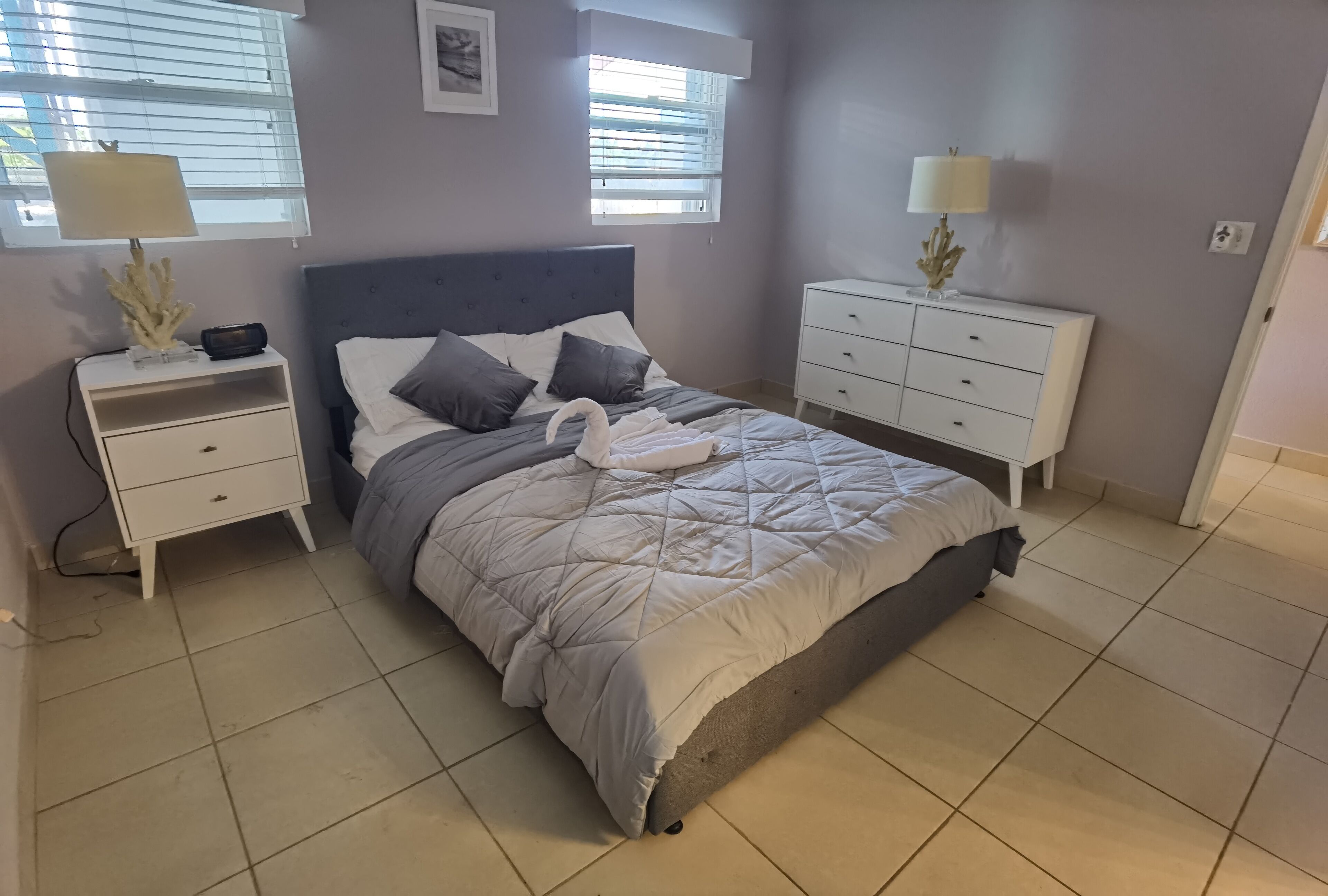 A bedroom with a bed, nightstands, a dresser, and two windows with blinds.