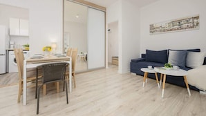 Uma sala de estar moderna com um sofá, uma mesa de jantar com cadeiras e uma área de cozinha visível através de uma porta deslizante.