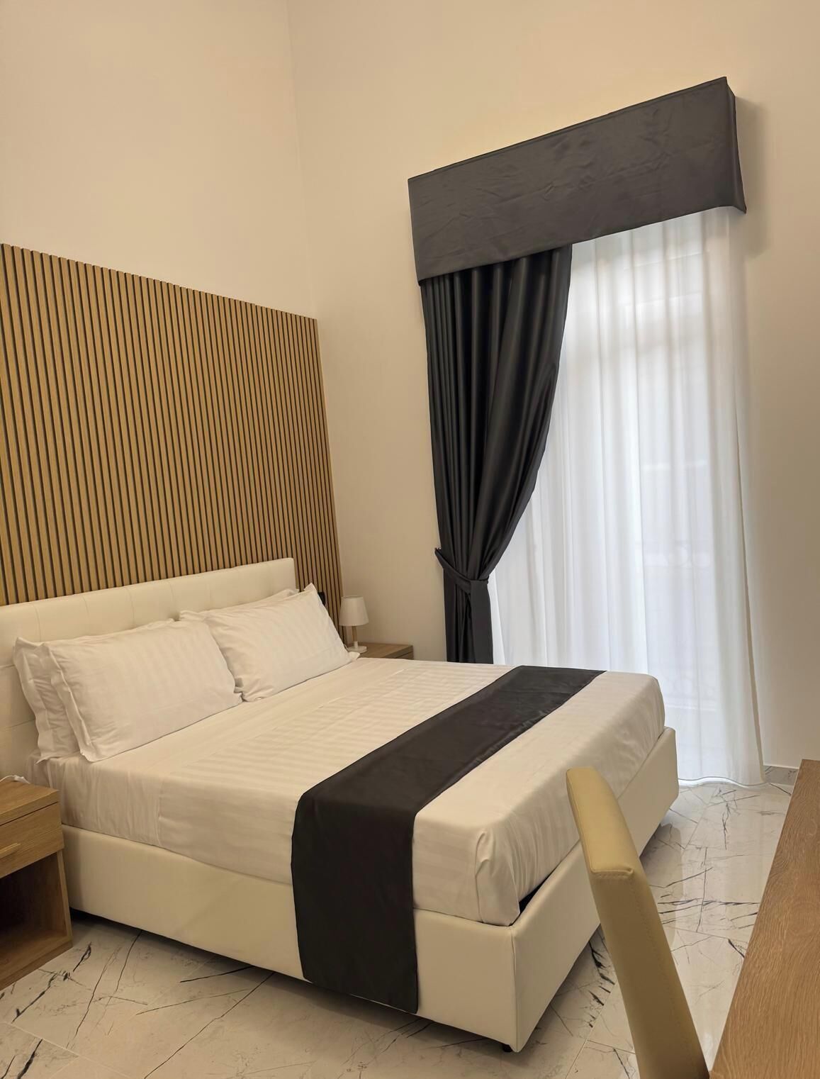 A hotel room with a bed, bedside tables, a nightstand, a window with curtains, and a headboard with vertical lines.