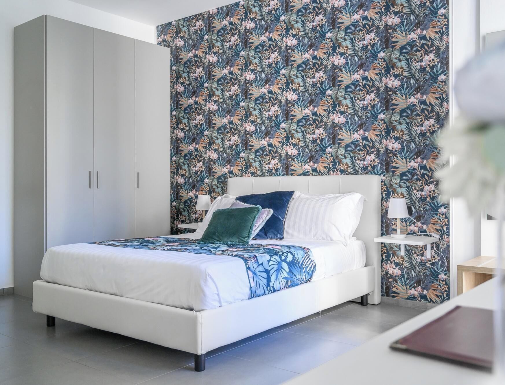 A modern bedroom with a white bed, floral wallpaper, and a nightstand with a lamp.