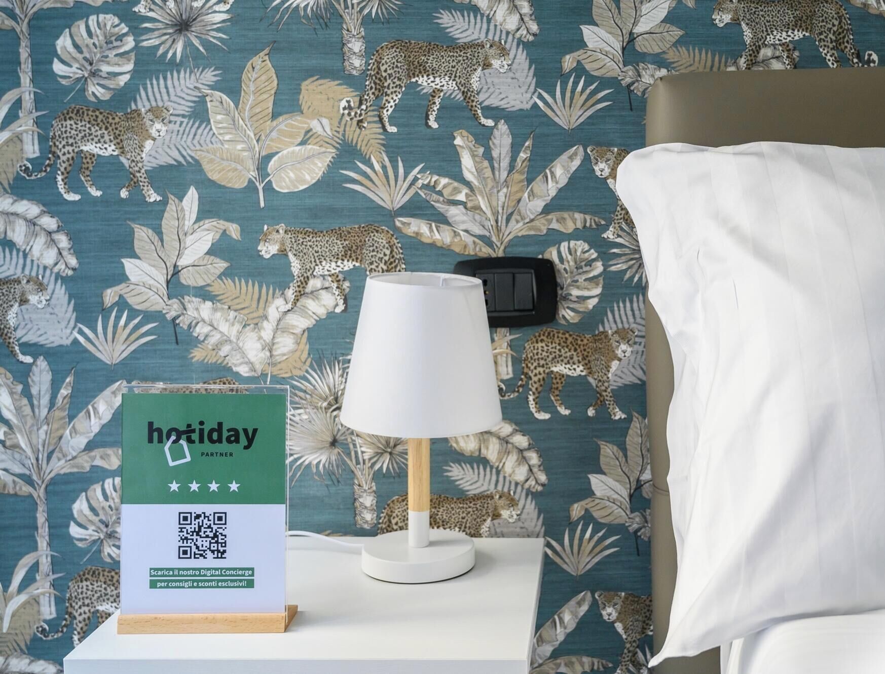 A hotel room with a leopard print wallpaper, a white bedside lamp, and a sign indicating a holiday partner.