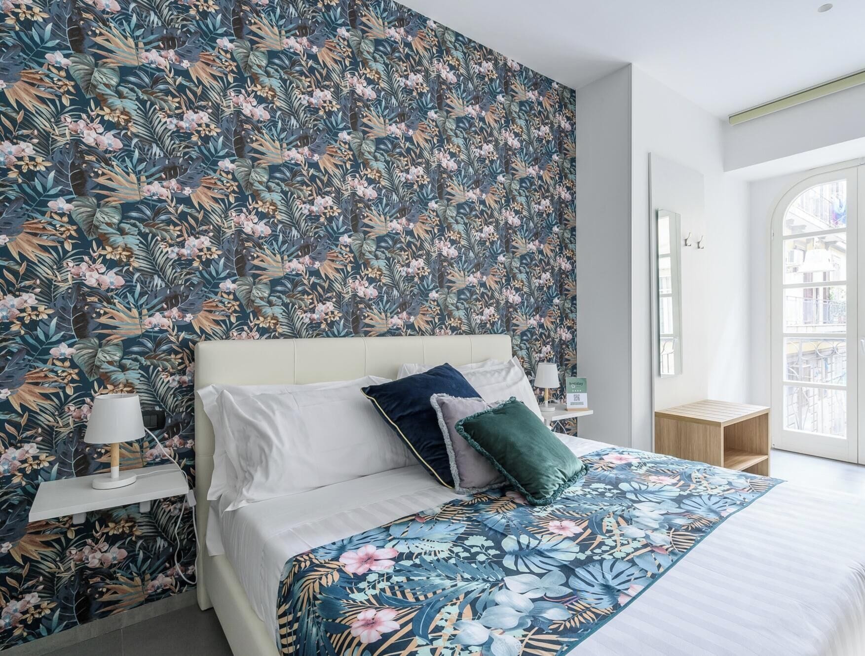 A bedroom with a floral wallpaper, a bed with white linens, and a nightstand with a lamp.