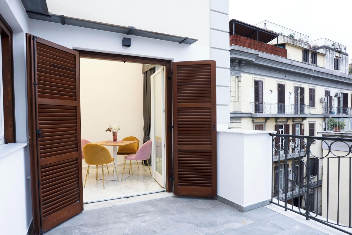 A balcony with a table and chairs, a door with wooden shutters, and a view of neighboring buildings.