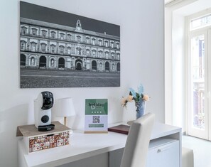 A hotel room with a desk, a coffee machine, a lamp, a vase with flowers, a framed picture of a building, and a QR code sign. - Hotiday Room Collection - Napoli Sant’Arcangelo  (Naples)