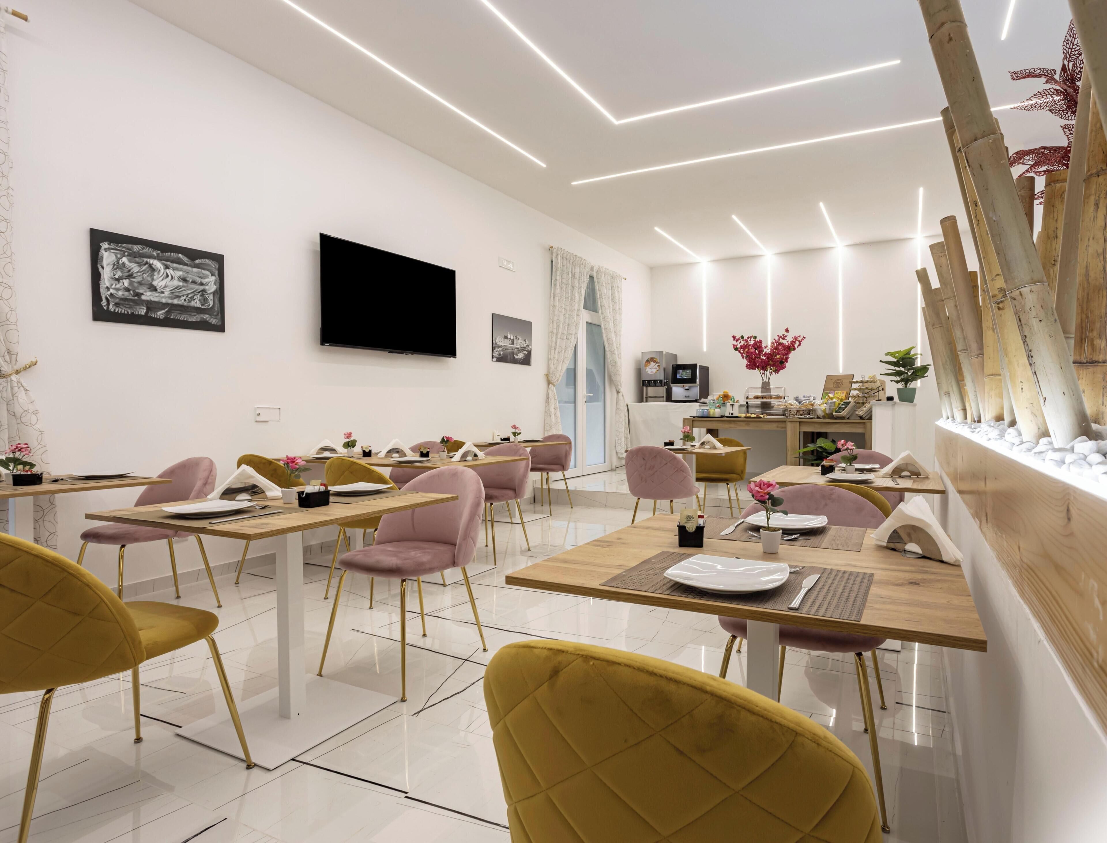 A modern dining area with multiple tables and chairs, a flat-screen TV mounted on the wall, and a kitchen area with a coffee machine.