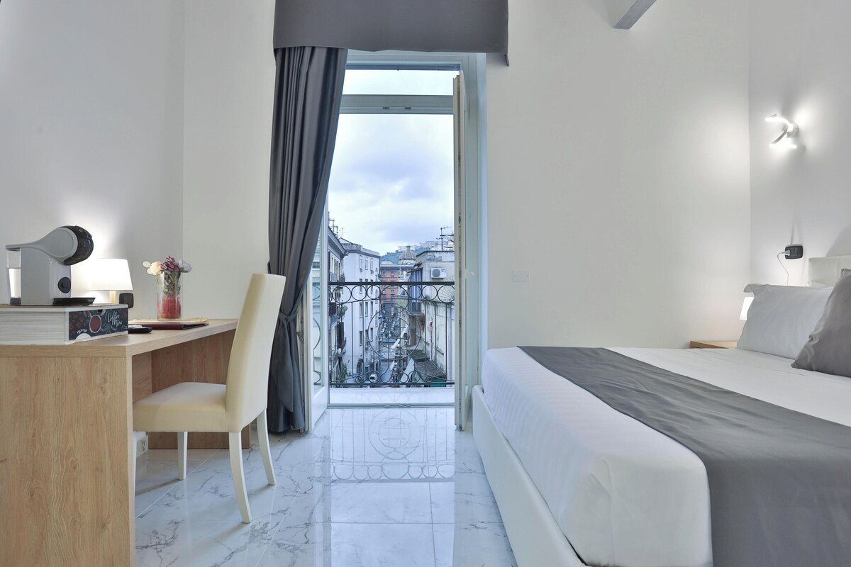 A modern hotel room with a bed, desk, chair, and a view of the city through a balcony door.