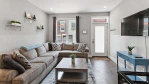 A modern living room with a sectional sofa, a coffee table, and a flat-screen TV. There are wall-mounted shelves with decorative items and a large window with curtains. - Skyline Best Roofdeck Views 2 Miles Penn Chop (Philadelphia)