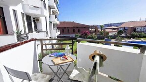 A balcony with a table and chairs, a hat, and a view of a swimming pool and buildings.