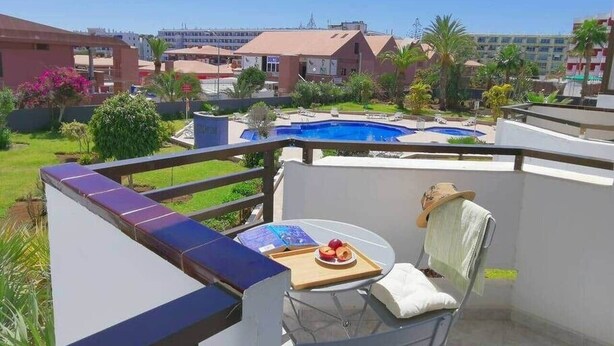 A balcony with a table, a chair, and a view of a swimming pool and buildings.