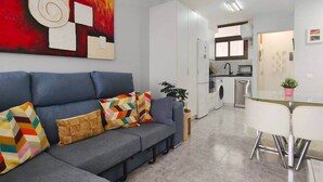 A modern living room with a grey sofa, colorful cushions, a large abstract painting, and a kitchen in the background.